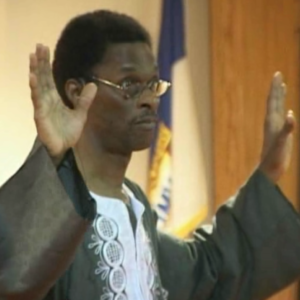 A man in glasses and a robe is holding his hands up.